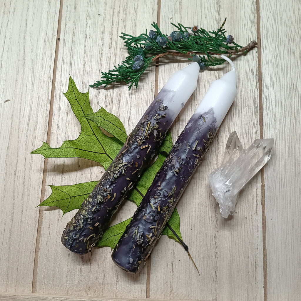 Two white candles with purple accents on a wooden surface with leaves and a crystal.