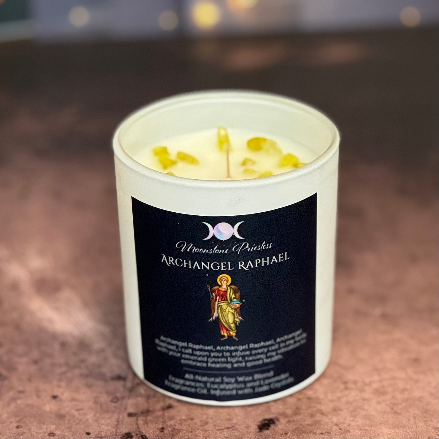 Candle in a white container with a black label featuring 'Archangel Raphael' on a blurred background.