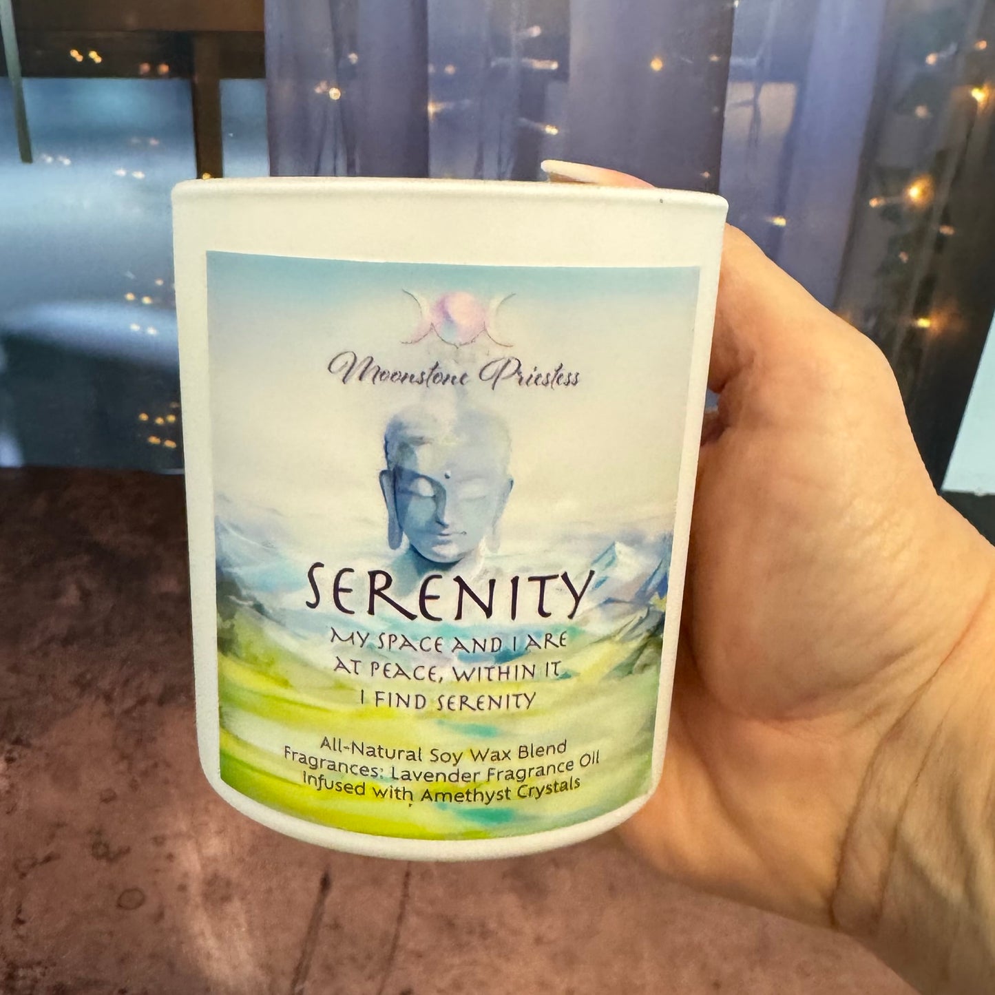 Hand holding a candle labeled 'Serenity' with a scenic design on the label.