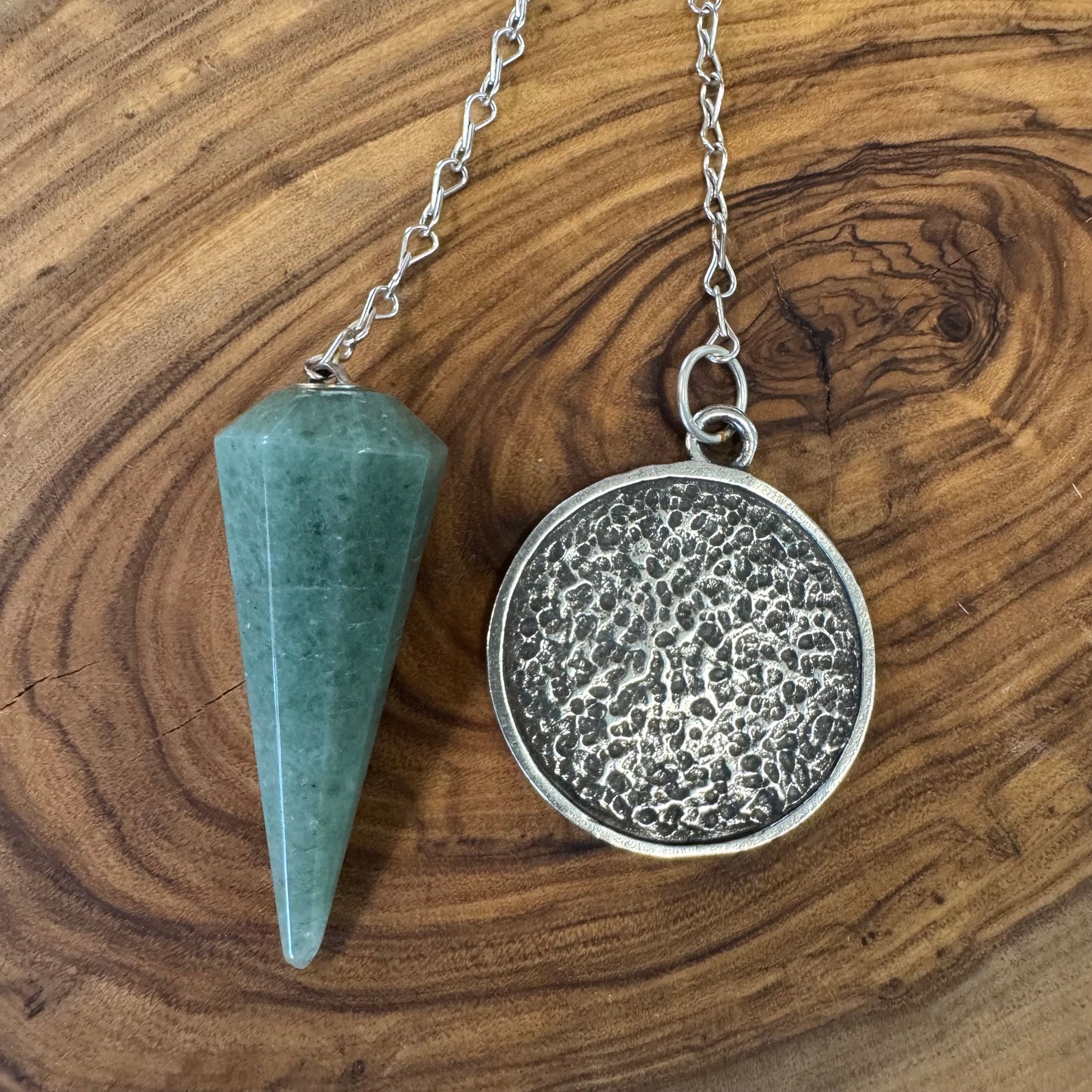 back of green aventurine pendulum on a wooden surface: a green stone cone and a silver round pendant with textured surface.