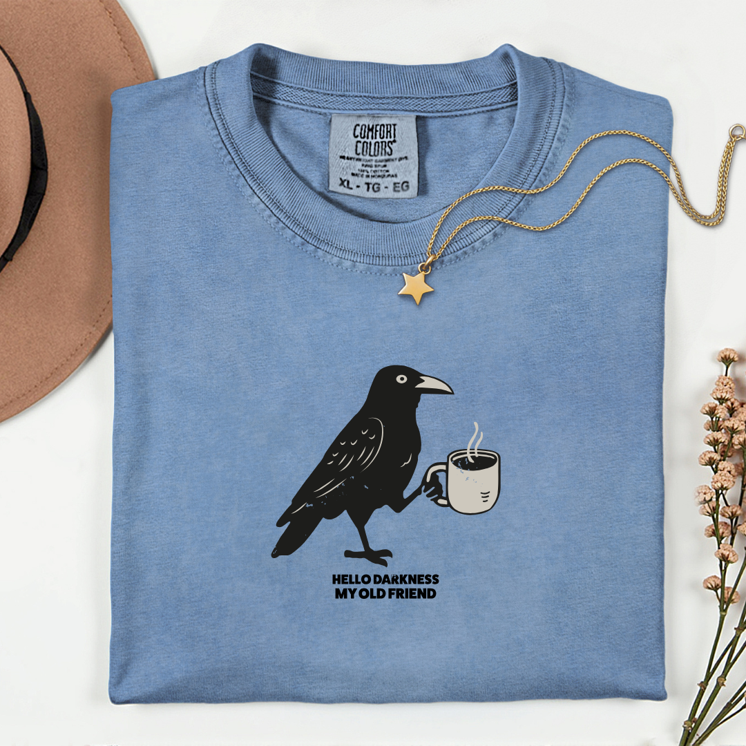 Folded blue t-shirt with a crow holding a coffee cup graphic and text on a white background.