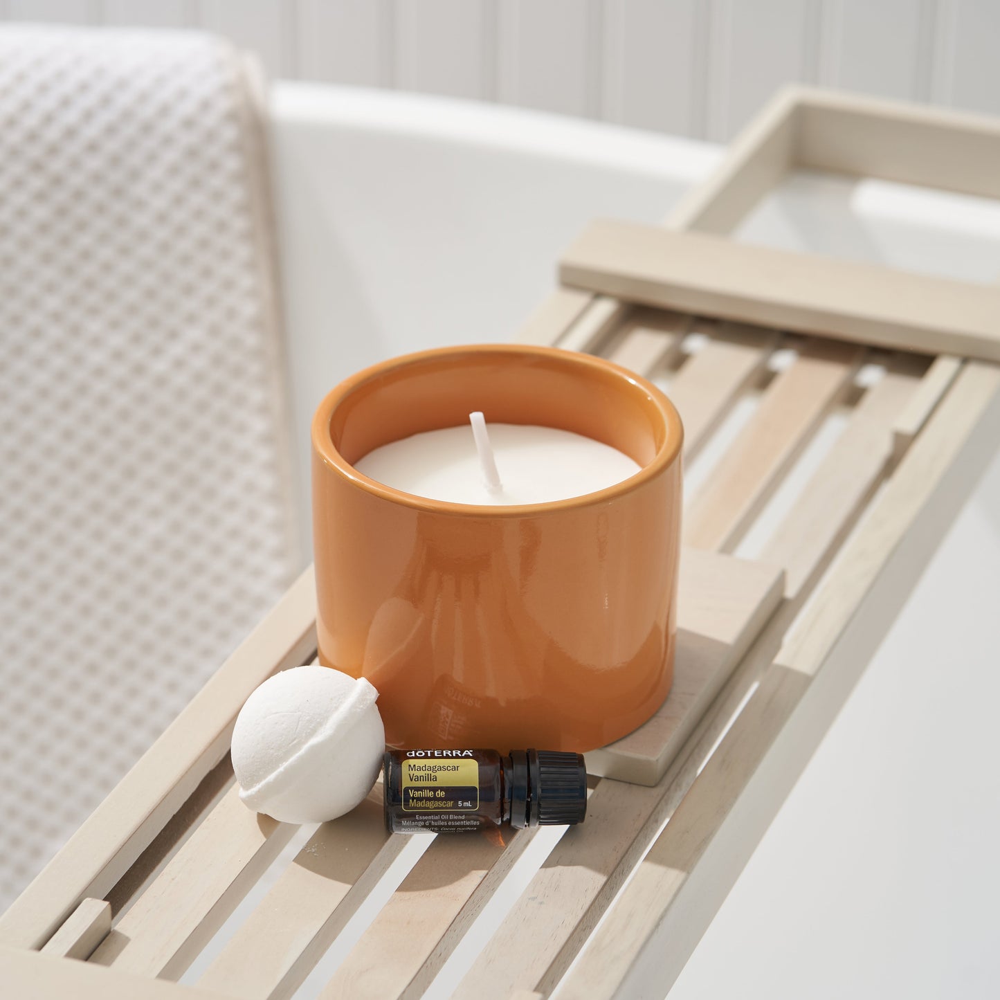 Candle in an orange holder with a small bottle and ball on a wooden tray against a white background