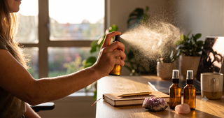Hand spritzing an aromatherapy intention spray at a desk beside crystals and a journal.