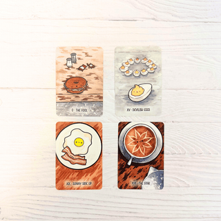 Four illustrated tarot-style cards arranged in a 2x2 grid on a light wood surface. The cards depict breakfast scenes: 'O.