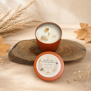 An orange metal candle tin with its lid placed in front, set on a rustic wood slice with dried leaves and wheat around it. The candle wax is visible in the open tin, and the lid label reads 'Smells Like a Waking Moon' and 'Moonphase Scent'.