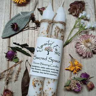 Two white house blessing candles tied with twine, wrapped in a white paper label reading 'Floral Fire Co. Sacred Space', arranged on a rustic wood surface with dried flowers and botanicals.