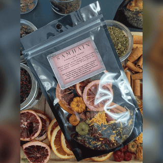 A clear stand-up pouch labeled 'SAMHAIN SUNFLOWER HERBS' filled with dried flowers, citrus slices, cinnamon, and other botanicals, photographed on a styled flat-lay with jars and dried fruit around it.
