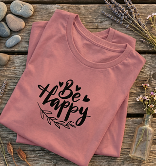 Folded Mauve Be Happy t-shirt with hand-lettered script, hearts, and a leaf design on a wooden surface with rocks and flowers