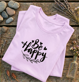 Folded Heather Prism Lilac Be Happy t-shirt with hand-lettered script, hearts, and a leaf design on a wooden surface with rocks and lavender