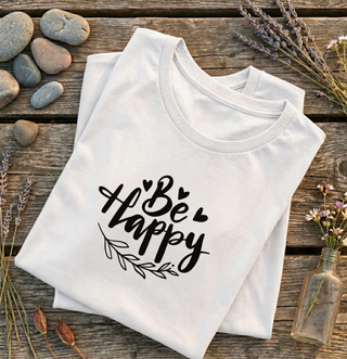 Folded Ash Be Happy t-shirt with hand-lettered script, hearts, and a leaf design on a wooden surface with lavender and stones