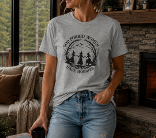 Athletic Heather God Forbid Women Have Hobbies t-shirt with a vintage moonlit forest design of two women dancing, worn in a cozy indoor setting