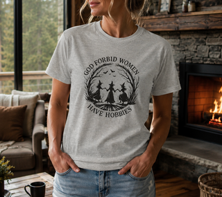 Athletic Heather God Forbid Women Have Hobbies t-shirt with a vintage moonlit forest design of two women dancing, worn in a cozy living room