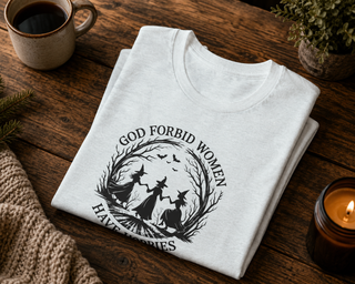 Folded white God Forbid Women Have Hobbies t-shirt with a vintage moonlit forest design on a wooden table with coffee and a candle