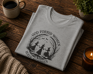 Folded Athletic Heather God Forbid Women Have Hobbies t-shirt with a vintage moonlit forest design on a wooden table with coffee and a candle