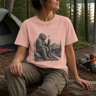 A woman sitting outdoors on a log in a forest setting, wearing a light pink crewneck t-shirt with a black graphic of Bigfoot sitting by a campfire. A green tent is visible in the background on the left.