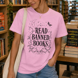 A woman in a bookstore wearing a light pink graphic t-shirt with the text 'READ BANNED BOOKS' in black, styled with a cream canvas shoulder bag and blue jeans.