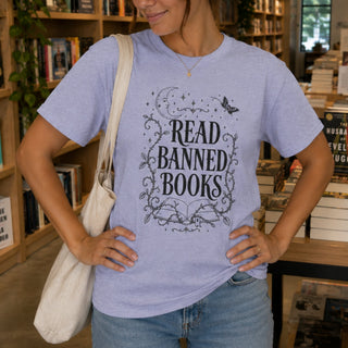 A woman in a bookstore wearing a light lavender heather t-shirt with a black graphic that reads 'READ BANNED BOOKS' surrounded by decorative moon, stars, and vines. She is carrying a cream canvas tote bag and wearing blue jeans.