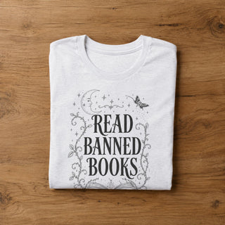 A folded white t-shirt with a black graphic print reading 'READ BANNED BOOKS' surrounded by a moon, stars, and butterfly design, laid flat on a wooden surface.