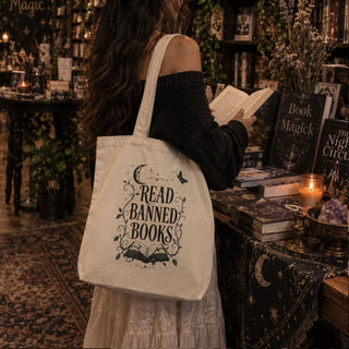 A woman in a cozy, dark-themed bookstore is shown from behind, wearing a large cream canvas tote bag with a black graphic that reads 'READ BANNED BOOKS' decorated with a crescent moon, open book, and stars. She is holding an open book next to tables filled with books and candles.