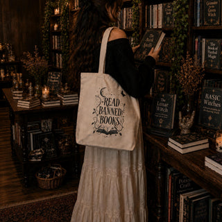 A woman in a black top and cream skirt stands in a cozy, dimly lit bookshop, carrying a cream canvas tote bag over her shoulder. The bag features a black graphic print that reads 'READ BANNED BOOKS' with illustrations of a crescent moon, stars, and an open book.