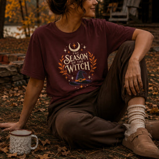 Cotton tee printed with a witch hat, crescent moon, stars, and orange laurel leaves beneath 'The Season of the Witch' lettering.