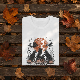 A folded white graphic T-shirt displayed on a rustic wooden bench surrounded by scattered autumn leaves. The shirt features a Halloween-themed illustration of two black cats sitting by a cauldron in front of an orange full moon, with stars and foliage details.