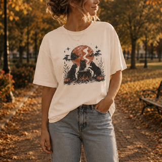 A woman wearing a cream-colored graphic t-shirt with a cat and moon design, paired with light-wash jeans, standing on an autumn leaf-covered path in a park.