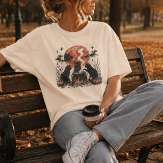 A woman sitting on a wooden park bench wearing an off-white graphic t-shirt with a black cat and campfire design, paired with light blue jeans and white sneakers, holding a coffee cup.