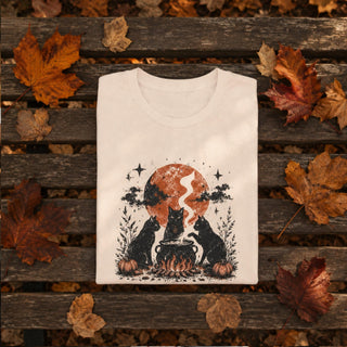 A folded off-white graphic T-shirt is placed on a weathered wooden bench surrounded by scattered autumn leaves. The shirt features a spooky Halloween-style print with two black cats sitting by a cauldron in front of an orange full moon and stars.
