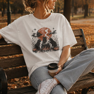 A woman sitting on a wooden park bench wearing a white graphic t-shirt with a Halloween-themed print of two black cats around a cauldron in front of a full moon. She is holding a coffee cup, with autumn leaves and trees in the background.