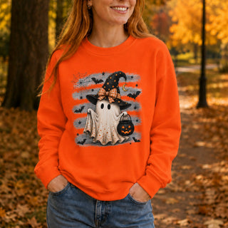 A woman wearing a bright orange crewneck sweatshirt with a Halloween graphic of a ghost in a witch hat holding a pumpkin lantern, photographed outdoors in an autumn park setting.