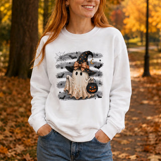 A woman wearing a white long-sleeve crewneck sweatshirt with a Halloween graphic of a ghost, witch hat, and jack-o'-lantern, set against an autumn park background.