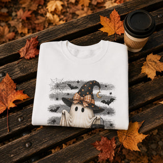 A folded white graphic T-shirt with a Halloween design featuring a ghost in a witch hat, placed on a dark wooden bench surrounded by autumn leaves and a takeaway coffee cup.