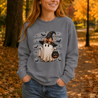 A woman wearing a gray sweatshirt with a cute Halloween graphic of a ghost in a witch hat, standing in an autumn park.