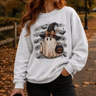 A woman wearing a light gray sweatshirt with a Halloween graphic of a ghost in a witch hat holding a pumpkin bucket, photographed outdoors on a leaf-covered path.
