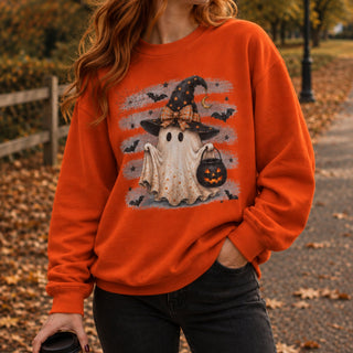A woman wearing an orange crewneck sweatshirt with a Halloween graphic of a ghost in a witch hat holding a pumpkin bucket, photographed outdoors on a path with fallen leaves.