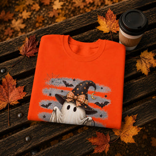 An orange crewneck sweatshirt with a Halloween graphic of a ghost in a witch hat, folded and styled on a dark wooden bench with autumn leaves and a coffee cup.