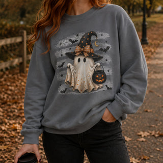 A woman wearing a gray crewneck sweatshirt with a Halloween graphic of a ghost in a witch hat holding a pumpkin bucket, photographed outdoors on a leaf-covered path.