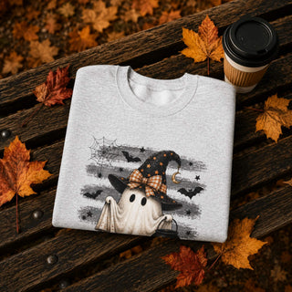A light gray folded sweatshirt with a Halloween-themed graphic print of a ghost wearing a witch hat, placed on a dark wooden bench surrounded by autumn leaves and a coffee cup.