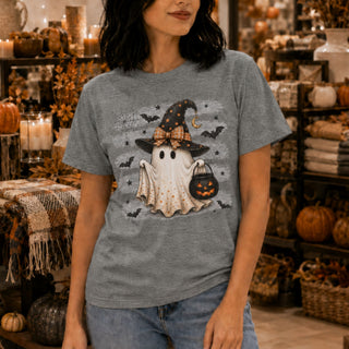 A woman wearing a heather gray graphic t-shirt with a cute Halloween ghost design, standing in a cozy, warmly lit retail shop filled with autumn decorations.