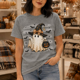 A woman wearing a heather gray graphic t-shirt with a cute Halloween ghost design, standing in a cozy, decorated retail store.