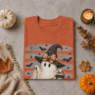 An orange heathered t-shirt with a Halloween graphic of a ghost wizard, styled in a flat lay with autumn props including pumpkins, a candle, and leaves on a gray concrete background.