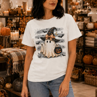 A woman wearing a white graphic t-shirt with a cute Halloween ghost design, standing in a cozy, autumn-themed boutique filled with pumpkins and candles.