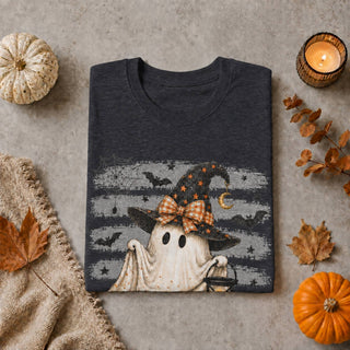 A folded dark charcoal gray t-shirt with a Halloween graphic print of a ghost wizard, styled in a flat-lay with autumn props including pumpkins, a candle, and leaves on a textured gray background.
