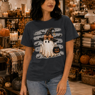 A woman wearing a dark navy blue graphic t-shirt with a Halloween design of a ghost in a witch hat holding a pumpkin lantern, set in a cozy, autumn-themed retail store.