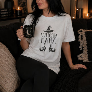 A woman sitting on a cozy couch wearing a white graphic t-shirt that reads 'WITCHY MAMA' with a witch hat and boots design, holding a black mug with a moon design.