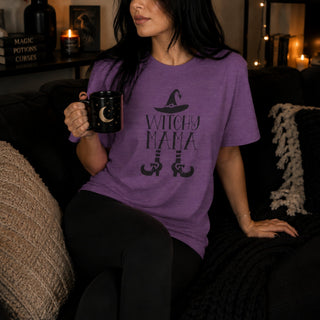 A woman sitting on a couch in a cozy room, wearing a purple graphic t-shirt that says 'WITCHY MAMA' with a witch hat and broomstick design. She is holding a black mug with a crescent moon.