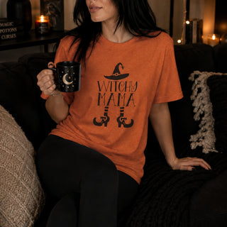 A woman wearing an orange crewneck t-shirt with a black 'Witchy Mama' graphic sits on a dark couch holding a black mug with a moon design.