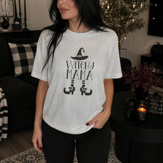 A woman wearing a white graphic t-shirt with a black 'WITCHY MAMA' design featuring a witch hat and legs, standing in a cozy, dimly lit living room.