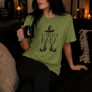 A woman sitting on a dark couch wearing a heather olive green t-shirt with a black graphic that reads 'WITCHY MAMA' with a witch hat and broomstick legs. She is holding a black mug with a crescent moon and stars, in a cozy, dimly lit room with candles and books in the background.